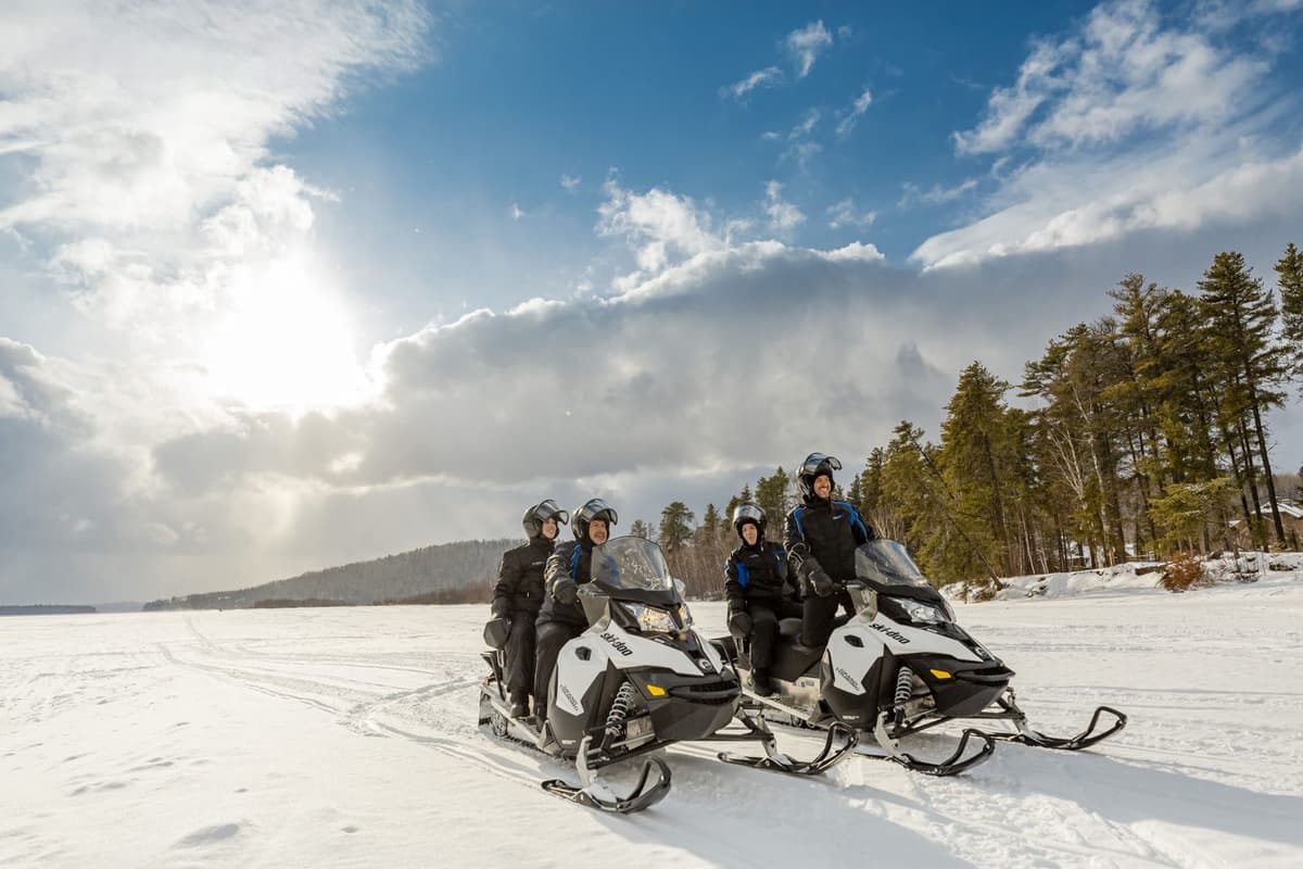 two snowmobiles and their four riders stopped under a sunny sky in the land of snowmobiling