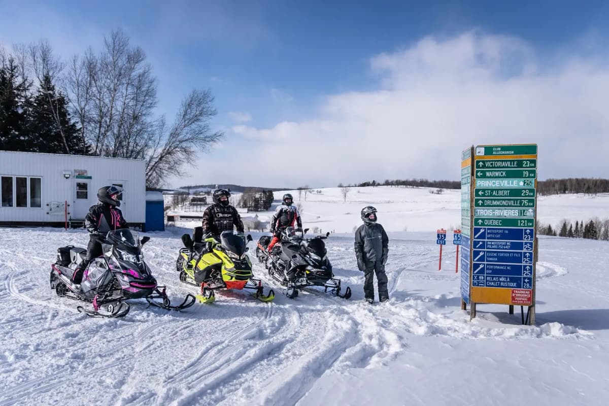 Balise ALT : Group of snowmobilers stopped near a direction sign on snowmobile trails in Québec.