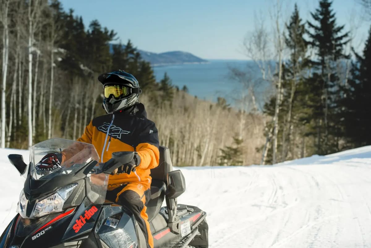 Snowmobiler in orange on a snow-covered trail with views of the mountains and sea.