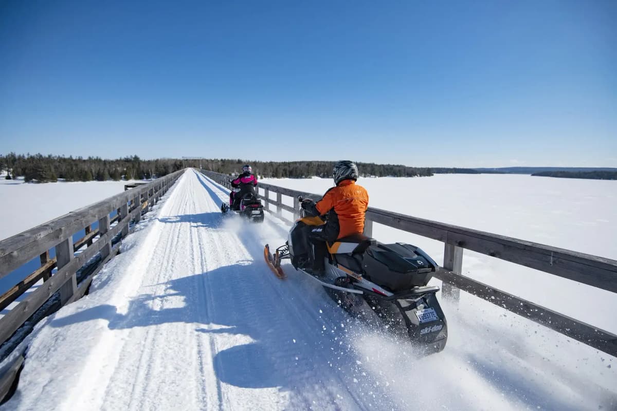 Snowmobilers crossing a snow-covered bridge over a frozen lake on snowmobile trails in Québec.