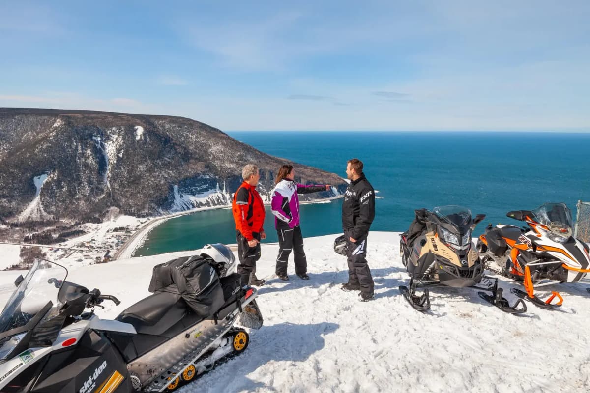 Snowmobilers chatting on top of a snow-covered cliff with sea views.