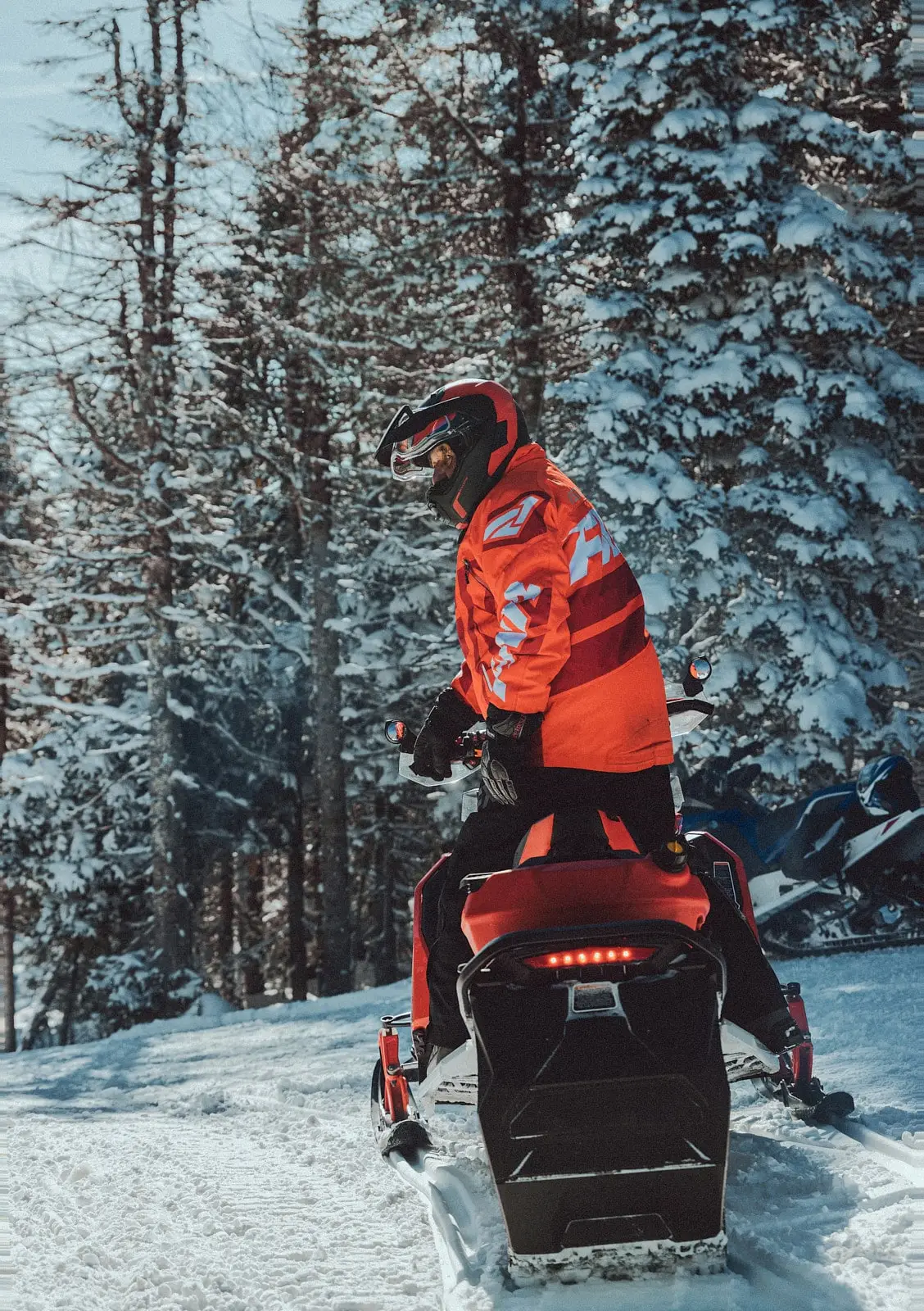 A snowmobiler in a red suit standing on their snowmobile, ready to traverse a snowy trail through a forest.