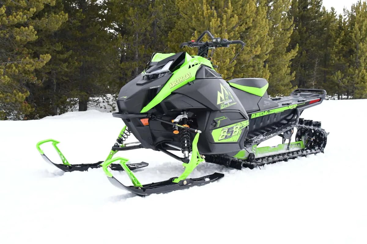 Close-up of a parked green M 858 Alpha One Sno Pro on a snowy terrain with a forest of evergreens behind it.
