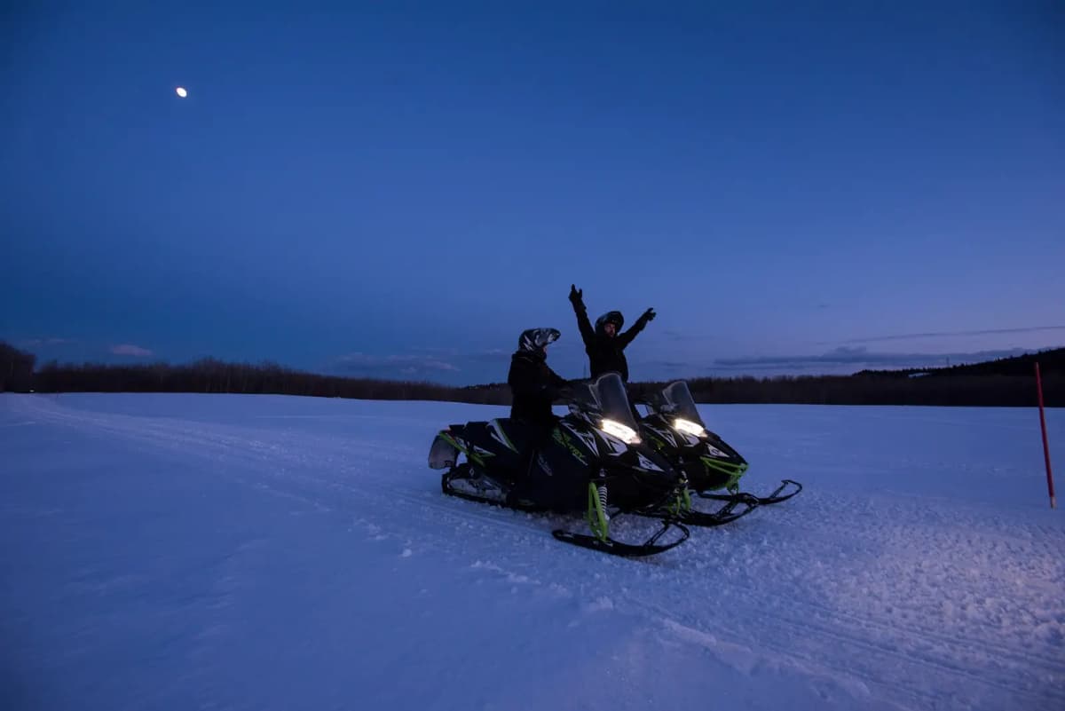 Two snowmobilers on the snowmobile trails in Abitibi-Témiscamingue, stop under a night sky, one raising his arms in sign of joy.