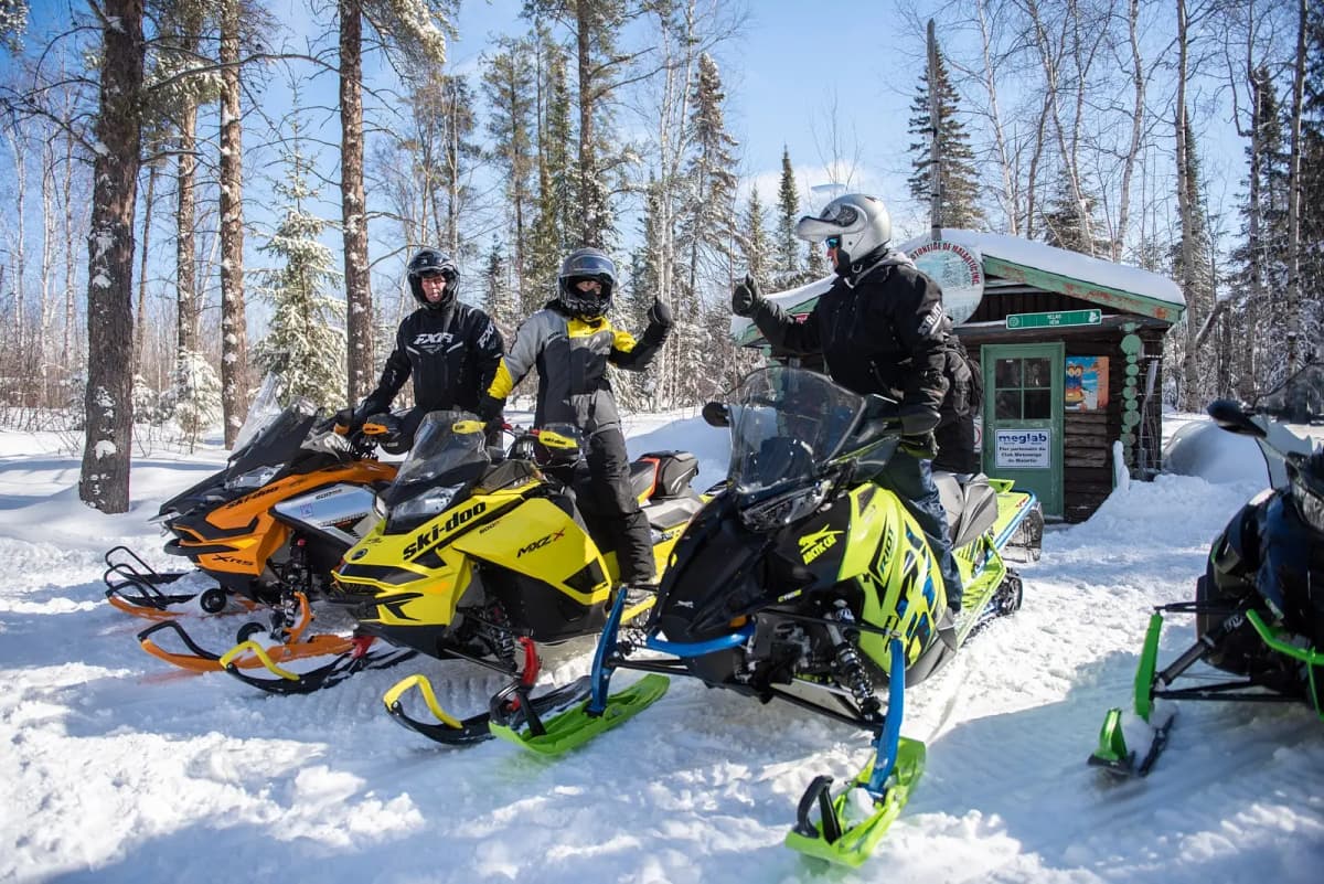 Three snowmobilers stopping near a wooden hut, swapping thumbs in a friendly atmosphere.