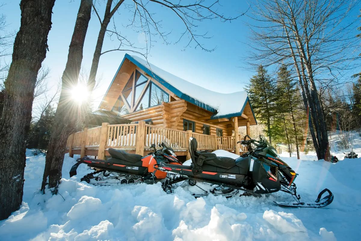 A cozy log cabin with a snow-covered roof, surrounded by trees and parked snowmobiles, illuminated by bright winter sunlight.
