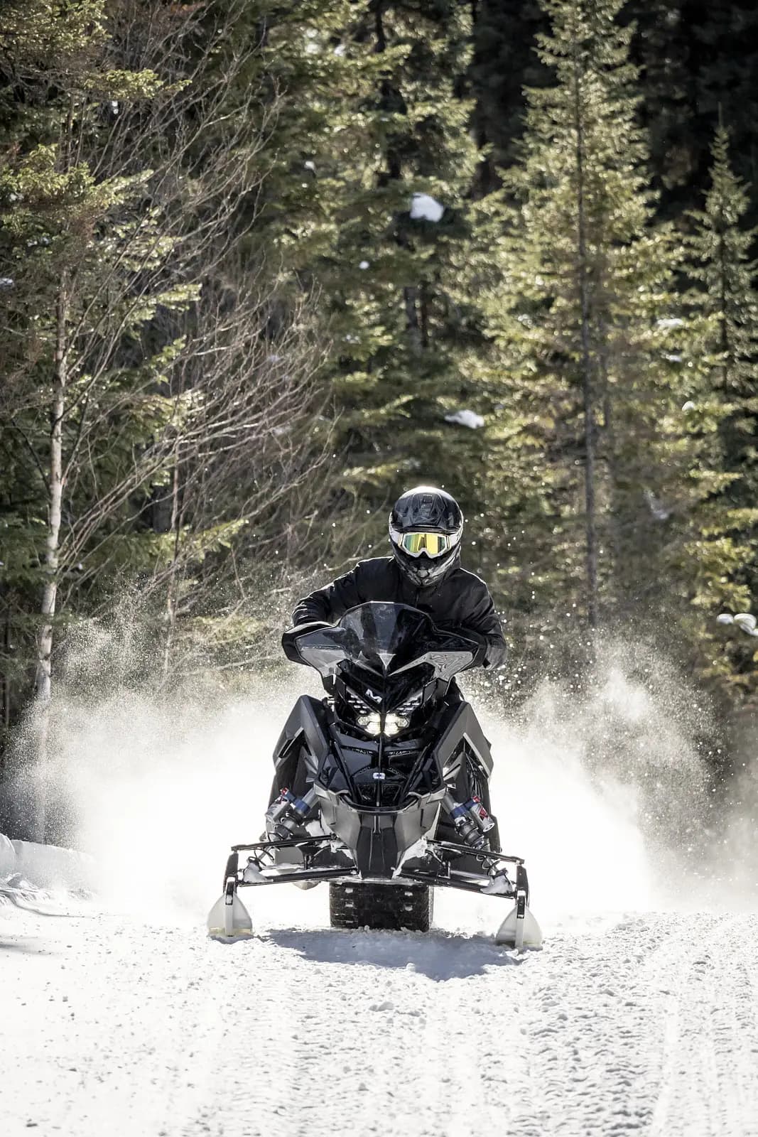A snowmobiler on a snowy trail surrounded by trees, kicking up a cloud of snow behind.