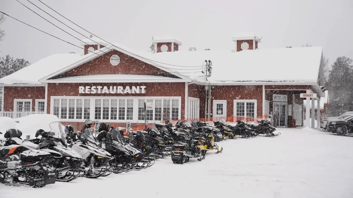 In front of a red wooden restaurant covered with snow in Lanaudière-Mauricie, a row of snowmobiles is parked while the snow falls slowly.