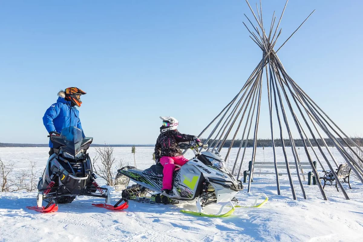 Two snowmobilers at a stop, one on a white Ski-Doo and the other on a black snowmobile, near a wooden tipi structure on a snowy landscape with a clear blue sky in the background.