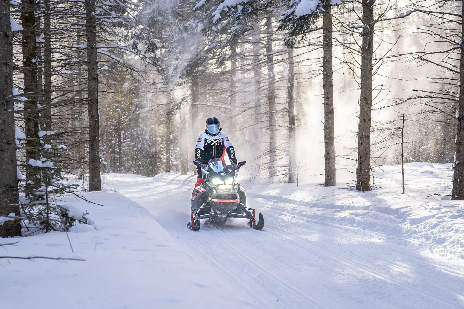 A snowmobile is travelling on a snowy trail surrounded by fir trees, with snow blown by the trees, in the Centre-du-Québec region.