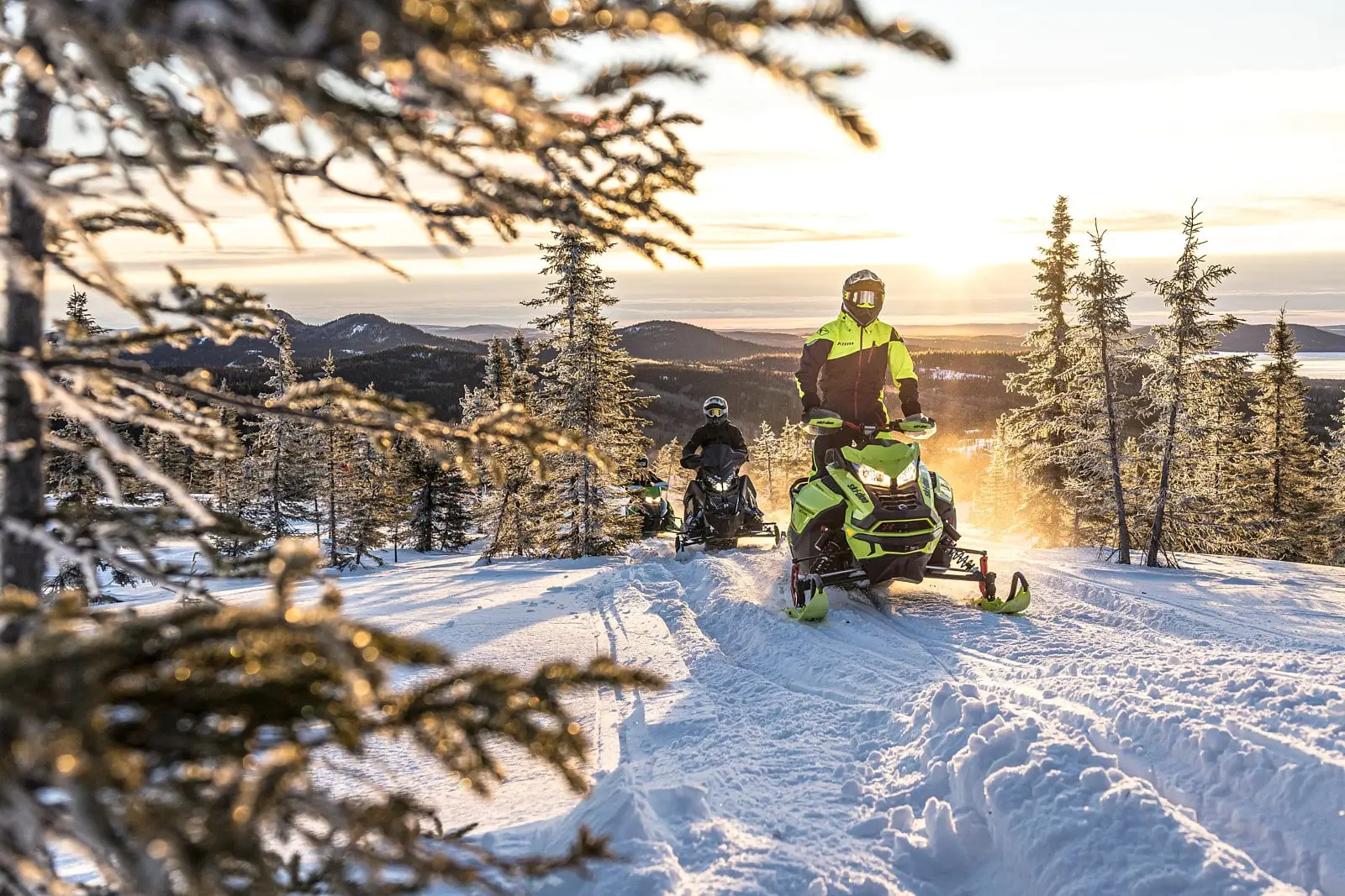 Two snowmobilers on a snowy trail in Eeyou Istchee Baie-James with a panoramic view of mountains and a sunset in the background.